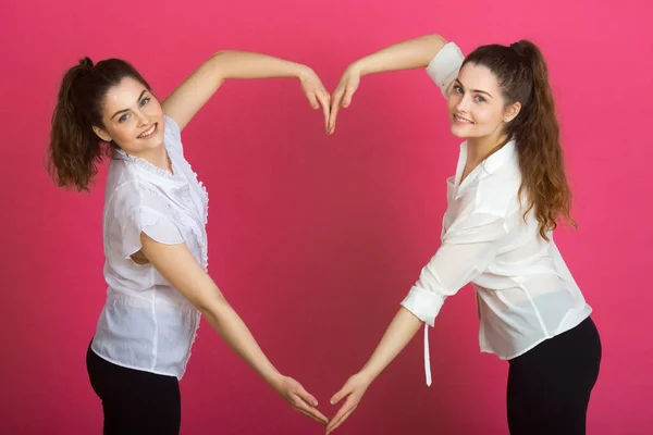 两个美丽的女孩姐妹双胞胎与微笑在粉红色的背景手势显示一个大的心脏失控 — 图库照片