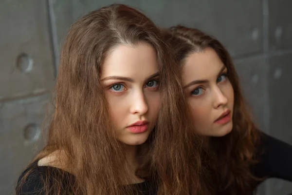 两个美丽的双胞胎姐妹的肖像与流动的头发对灰色的墙壁在内部 — 图库照片