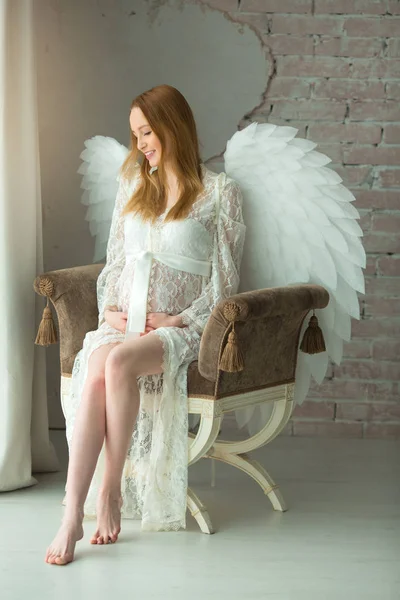 年轻美丽的孕妇在白色花边礼服与翅膀坐在室内 — 图库照片