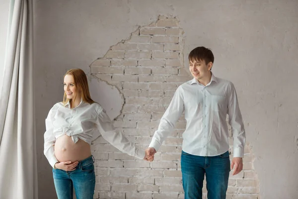 美しい夫と妊娠中の妻のジーンズと白いシャツ白い壁近くに立っています — ストック写真