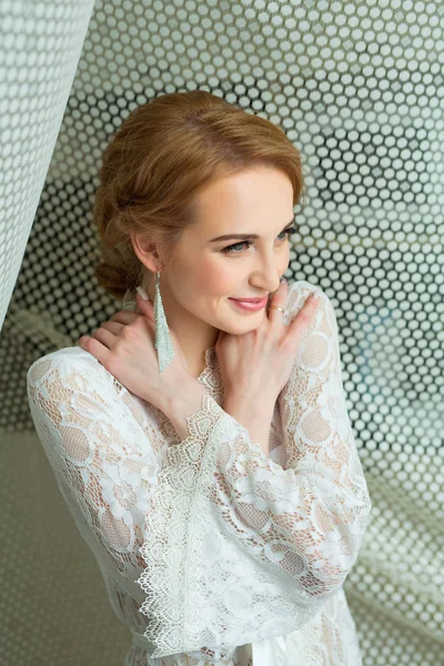 美丽的年轻新娘在白色花边敷料站在窗口附近 — 图库照片