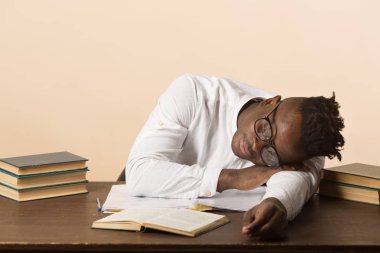 Gözlüklü ve beyaz gömlekli yakışıklı Afrikalı bir adam bej arka planda dizüstü bilgisayarı olan bir masada oturuyor.