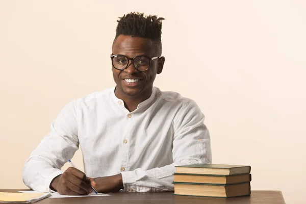 handsome young african man in glasses and a white shirt sits at a table with books on a beige background