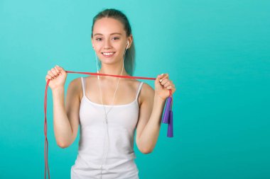 Mavi arka planda spor giyimli genç ve güzel bir kadın. Elinde ip atlıyor.