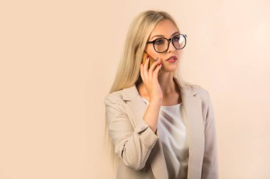 Bej arka planda takım elbiseli güzel genç bir kadın. Elinde telefon var.