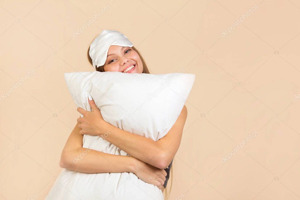 beautiful young woman on a beige background in a sleep mask with a pillow in her hands