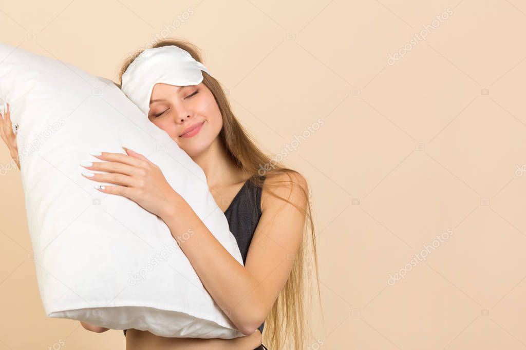 beautiful young woman on a beige background in a sleep mask with a pillow in her hands