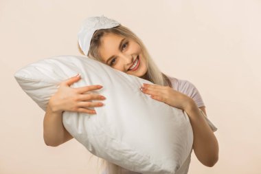 Bej bir arka planda uyku maskesi takmış, elinde yastıkla güzel bir genç kadın.