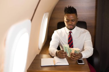 Elinde dolarlarla, beyaz gömlekli, uçak koltuğunda oturan yakışıklı bir Afrikalı.