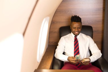 Elinde dolarlarla, beyaz gömlekli, uçak koltuğunda oturan yakışıklı bir Afrikalı.