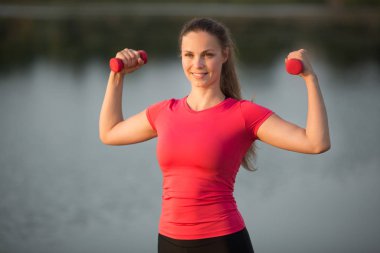 Spor giyim sektöründe genç ve güzel bir kadın gölün kenarındaki bir parkta elinde dambıllarla çalışıyor.