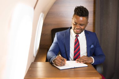 Takım elbiseli yakışıklı Afrikalı genç adam özel jet kabininde elinde not defteriyle