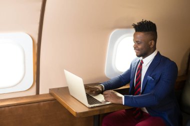 Özel jet kabininde dizüstü bilgisayarlı, takım elbiseli yakışıklı Afrikalı genç adam.