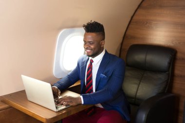 Özel jet kabininde dizüstü bilgisayarlı, takım elbiseli yakışıklı Afrikalı genç adam.