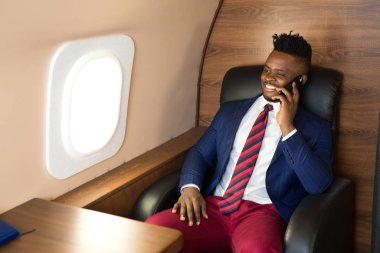 Elinde telefonla özel bir jetin kabininde takım elbiseli yakışıklı Afrikalı genç bir adam.