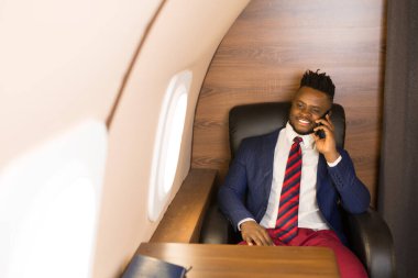 Elinde telefonla özel bir jetin kabininde takım elbiseli yakışıklı Afrikalı genç bir adam.