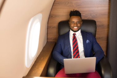 Özel jet kabininde dizüstü bilgisayarlı, takım elbiseli yakışıklı Afrikalı genç adam.