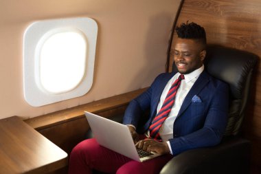 Özel jet kabininde dizüstü bilgisayarlı, takım elbiseli yakışıklı Afrikalı genç adam.