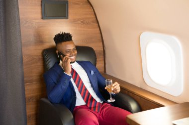 Elinde telefonla özel bir jetin kabininde takım elbiseli yakışıklı Afrikalı genç bir adam.