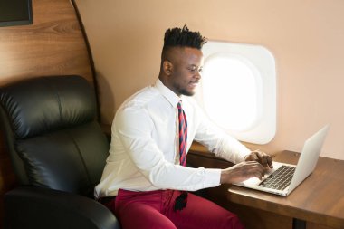 Özel jet kabininde dizüstü bilgisayarlı beyaz gömlekli yakışıklı Afrikalı genç adam.