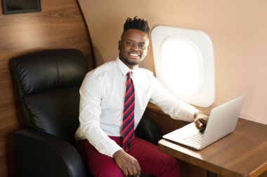 Özel jet kabininde dizüstü bilgisayarlı beyaz gömlekli yakışıklı Afrikalı genç adam.
