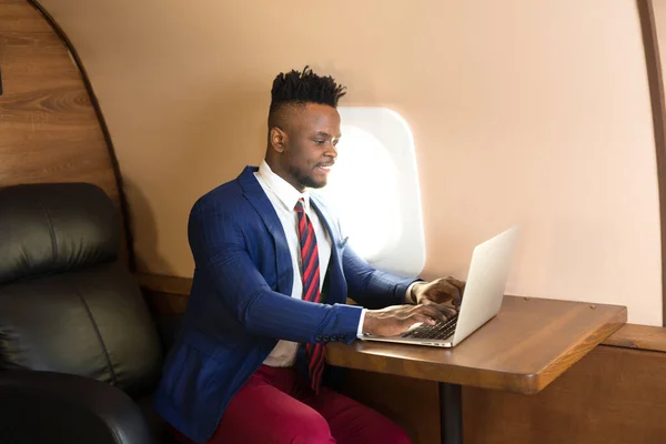 handsome african young man in suit in private jet cabin with laptop