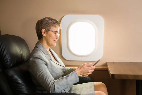 adult woman with a short haircut in a suit sits in the chair of a private plane with a phone in her hands