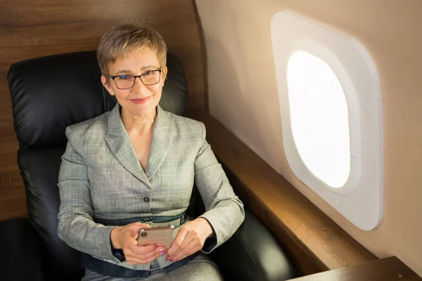 Kısa saçlı, takım elbiseli yetişkin bir kadın elinde telefonuyla özel bir uçağın koltuğunda oturuyor.
