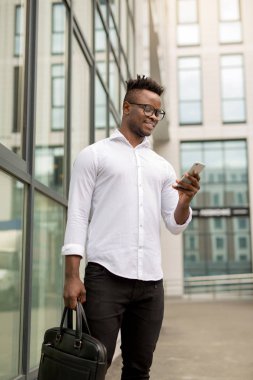 Beyaz tişörtlü yakışıklı Afrikalı bir adam. Elinde telefon, cam bir binanın yanında.