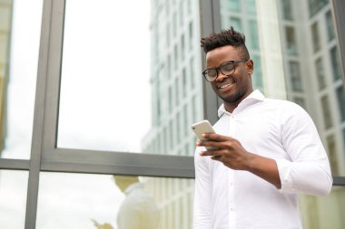 Beyaz tişörtlü yakışıklı Afrikalı bir adam. Elinde telefon, cam bir binanın yanında.