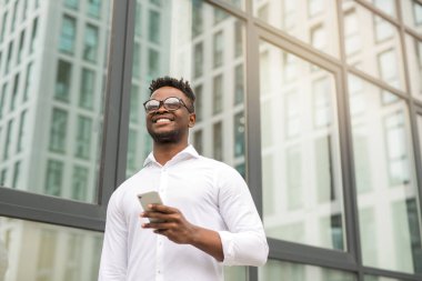 Beyaz tişörtlü yakışıklı Afrikalı bir adam. Elinde telefon, cam bir binanın yanında.