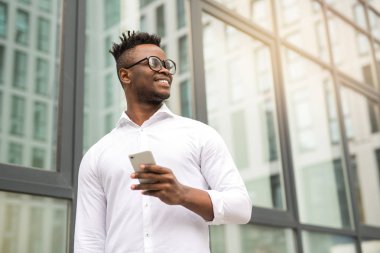 Beyaz tişörtlü yakışıklı Afrikalı bir adam. Elinde telefon, cam bir binanın yanında.