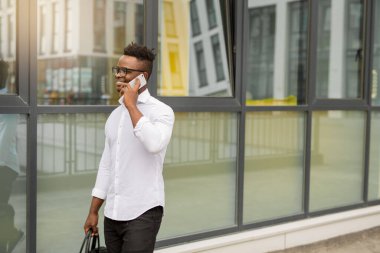 Beyaz tişörtlü yakışıklı Afrikalı bir adam. Elinde telefon, cam bir binanın yanında.