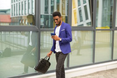 Takım elbiseli yakışıklı, genç Afrikalı bir adam. Elinde telefon, cam bir binanın yanında.