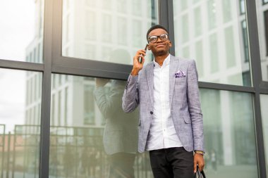 Takım elbiseli yakışıklı, genç Afrikalı bir adam. Elinde telefon, cam bir binanın yanında.