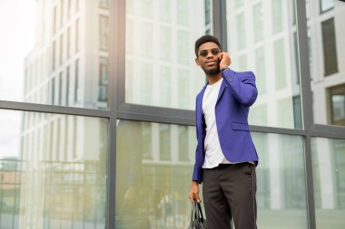 Takım elbiseli yakışıklı, genç Afrikalı bir adam. Elinde telefon, cam bir binanın yanında.