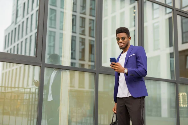 handsome young african man in a suit with a phone in his hands near a glass building