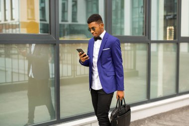 Takım elbiseli yakışıklı, genç Afrikalı bir adam. Elinde telefon, cam bir binanın yanında.
