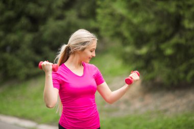 Spor pembesi tişörtlü, elinde dambıllar, yaz parkında.