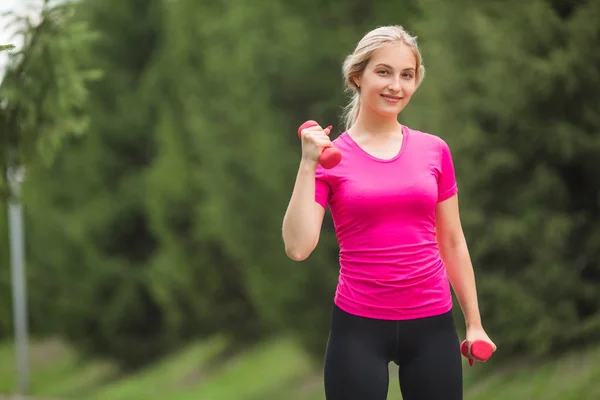 beautiful young woman in sports pink t-shirt with dumbbells in hands in the summer park