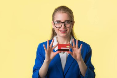 Sarı arka planda mavi ceketli, elinde kredi kartıyla güzel bir genç kadın.