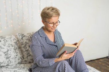 Yatak odasında pijamalı yaşlı bir kadın kitap okuyor.