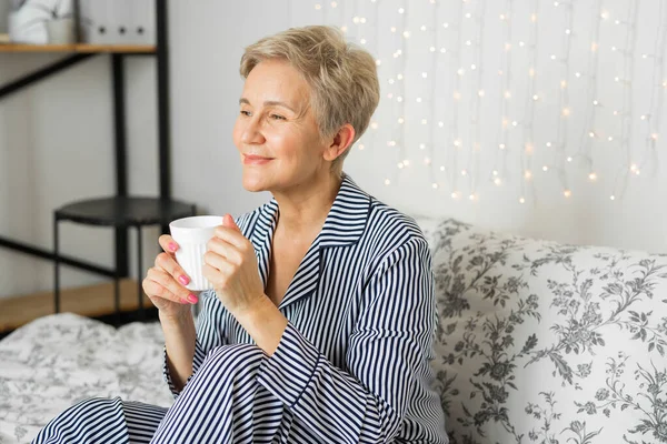 Yatak odasında pijamalı yaşlı bir kadın elinde kupayla yatıyor.