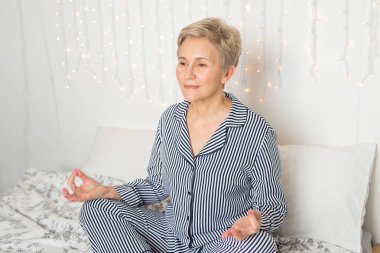 Güzel, yaşlı bir kadın, pijamalarıyla yoga pozisyonunda yatakta.
