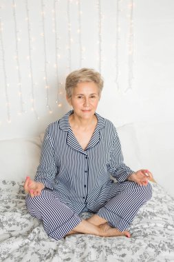 Güzel, yaşlı bir kadın, pijamalarıyla yoga pozisyonunda yatakta.