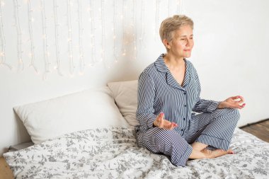 Güzel, yaşlı bir kadın, pijamalarıyla yoga pozisyonunda yatakta.