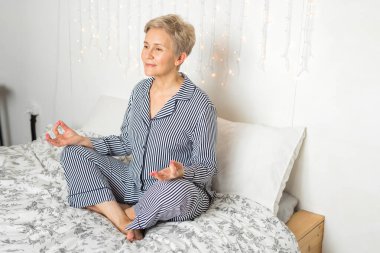 Güzel, yaşlı bir kadın, pijamalarıyla yoga pozisyonunda yatakta.