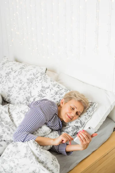 beautiful elderly woman in pajamas on the bed under the covers with a phone