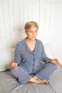 Güzel, yaşlı bir kadın, pijamalarıyla yoga pozisyonunda yatakta.
