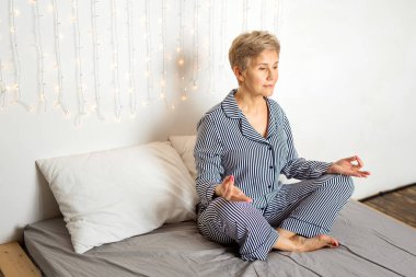 Güzel, yaşlı bir kadın, pijamalarıyla yoga pozisyonunda yatakta.